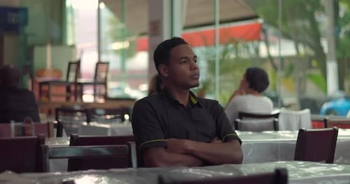 Young male restaurant employee seated alone with arms crossed, gazing forward with serious and