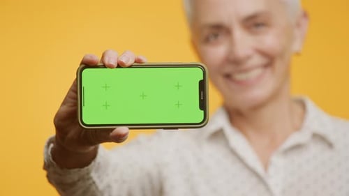 Mature Woman Holding Green Screen Smartphone on Yellow