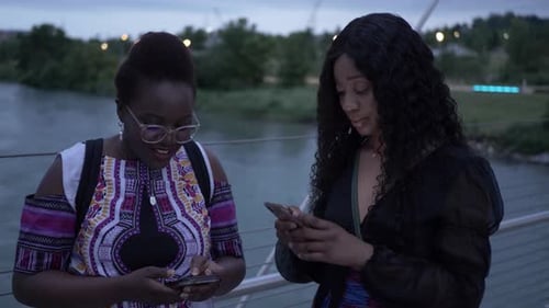 Young women friends texting together on bridge over river at night in the city