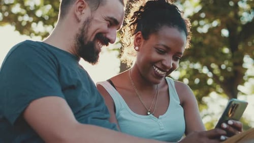 Happy smiling interracial couple watching video on mobile phone while sitting on bench
