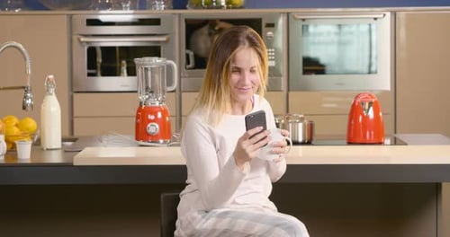 Woman in Pajamas Uses Phone, Holds Mug in Kitchen