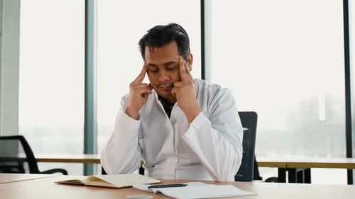 Stressed Frustrated Indian Doctor with Headache Sitting Alone at Work Desk in Hospital Sad Young Gp