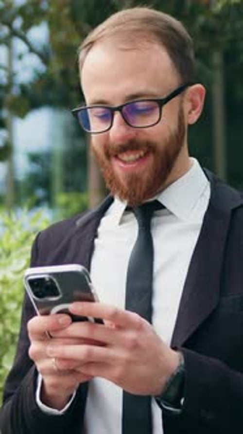 Young Smiling Businessman is Using a Smartphone While Sitting on a Bench Near an Office Building