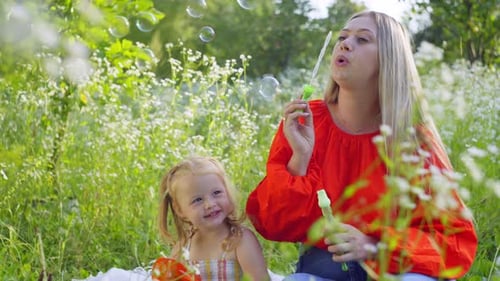 Mother Plays with Her Daughter Sitting in Grass in Field Blowing Soap Bubbles