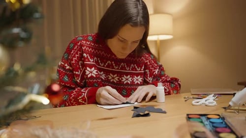 A Woman in a Red Festive Sweater Works on a Craft Project at a Table Adorned with Holiday Supplies.
