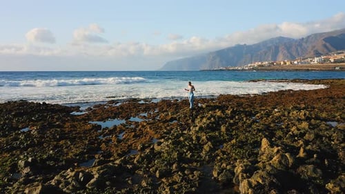 Girl Traveler Explores Walking on Rocky Volcanic Beach with Wide Foaming Waves and Huge Cliffs on