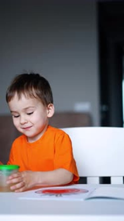 Concentrated three-year-old toddler wearing orange t-shirt moves the brush by the paper.