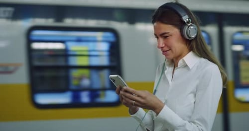 Woman Using Headphones and Smartphone to Listen to Music at Subway Station While Waiting for P