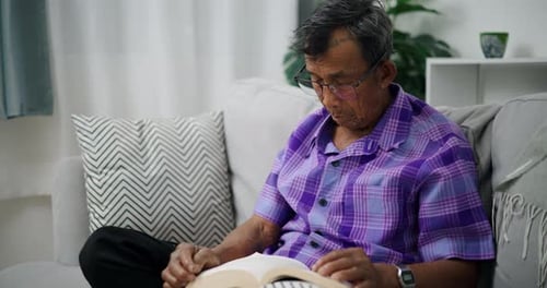 Senior man wearing glasses sits reading a book relaxing on a sofa at home.