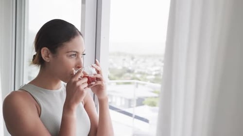 Thoughtful biracial woman looking through window drinking tea at home, copy space, slow motion