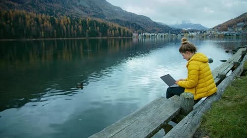 Young woman in bright yellow jacket work on side of lake on her laptop. Female freelancer