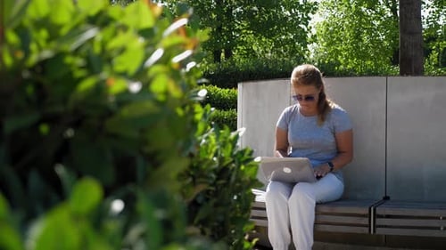 A Woman in Sunglasses is Sitting on a Wooden Bench with a Laptop in a Park