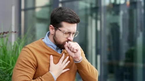 A sick businessman coughing sitting on a bench on the street near office building. Upset male worker