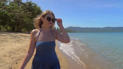 Closeup of Young European Girl in Sunglasses and Blue Dress Walking on the Sandy Beach Against the