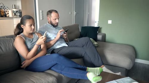 Father and teen daughter using phones on sofa