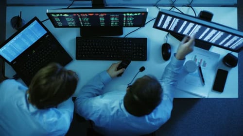 Ansicht von oben auf zwei Börsenmakler, die Marktzahlen suchen und analysieren. Vor ihnen zwei Monitore mit S