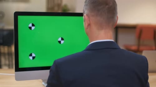 A man in a business suit works at a computer with a large monitor, green screen