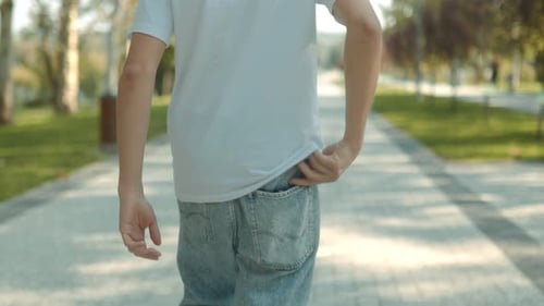 Rear View of a Young Man Taking Phone Out While Walking in Park on a Sunny Day