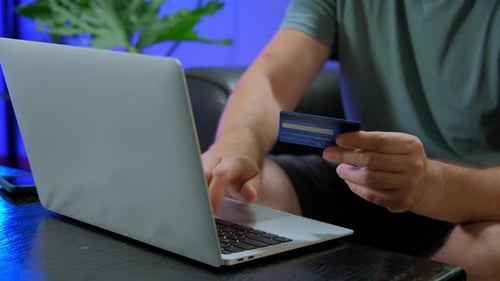 Mann sitzt auf der Couch im Wohnzimmer mit Karte und benutzt Laptop-Computer beim Online-Shopping