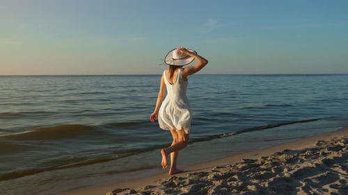 Glückliche junge Frau in weichem Kleid und stilvollem Hut läuft den Strand entlang