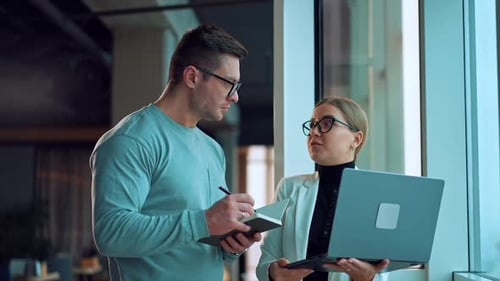 People working together in office communicating standing at the window. Lady holding a laptop