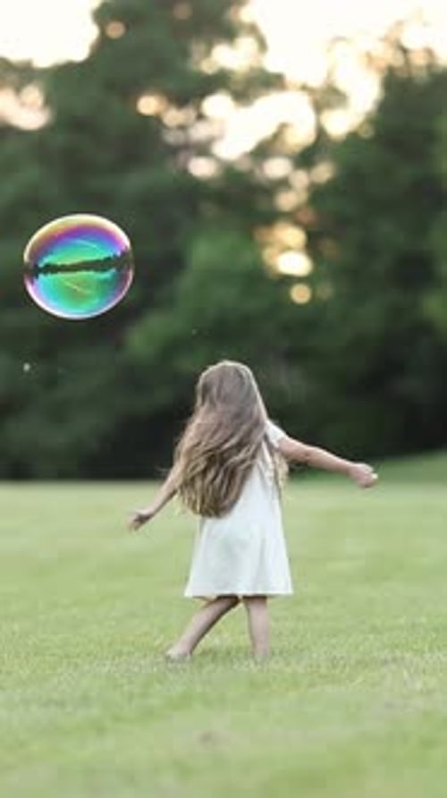 Young Girl in White Dress Reaching Up to Catch a Soap Bubble While Playing on a Grassy Field