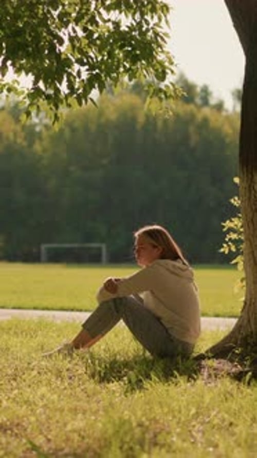 Woman Sitting Thoughtfully By Tree in Sunlit Park Surrounded By Greenery