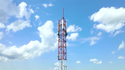 Cell phone network tower with 5 g antennas in red and white with blue sky and white clouds in the ba