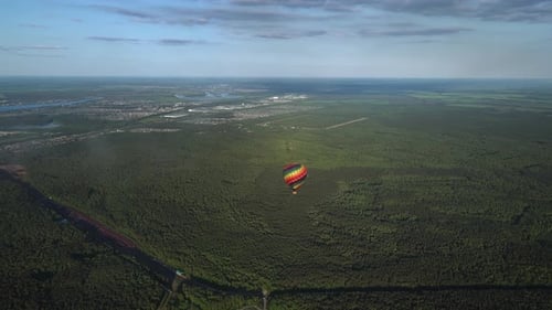 Lonely Colorful Hot Air Balloon is Flying Over Green Forest at Clear Sky with Town on Background