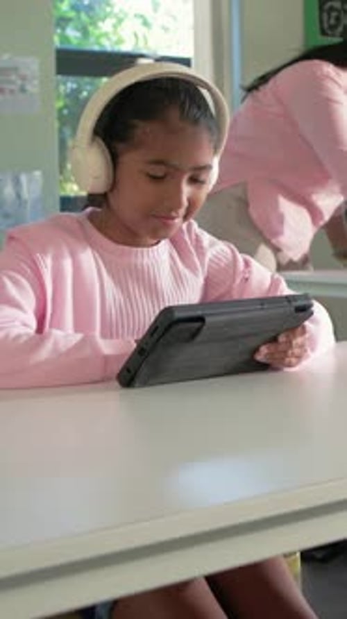 Vertical video: Pausing, kid in pink top and cream headset tapping tablet at school desk, learning