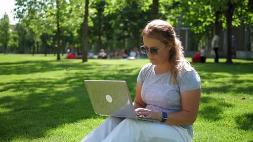A Focused Woman in Sunglasses is Working with a Laptop in a Summer Park