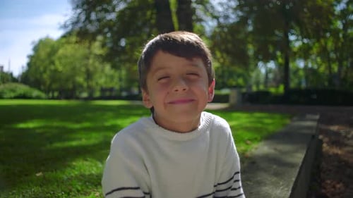 Young boy closing eyes with big smile outdoors in park, wearing striped sweater and radiating joy
