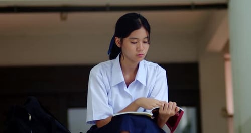 A cute Thai or Asian high school girl in a white school uniform reading a book for exam preparation,