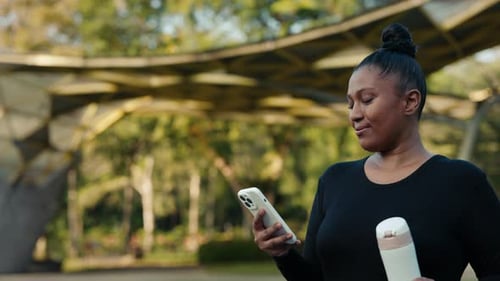 Woman in Sportswear Walking Down Street After Run Holding Sports Water Bottle Using Telephone