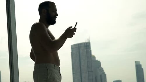 Silhouette of Man Texting on Smartphone Standing by Window at Home 30s