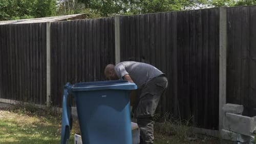Adult UK Asian Male Sorting And Ripping Cardboard Beside Blue Recycling Bin In Garden. Locked Off