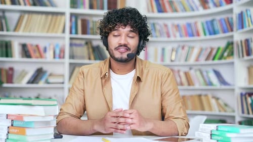 Young happy college teacher in headset conducts online video call training in campus library space.