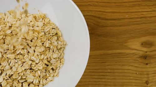 Oatmeal is poured into a white ceramic bowl against a wooden board. Top view. Slow motion.