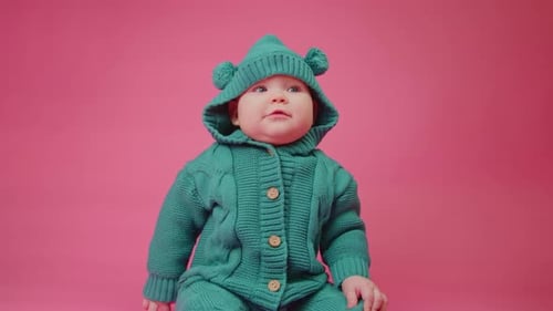 Portrait of a Cute Baby in a Warm Knitted Jumpsuit with a Hood on a Pink Background