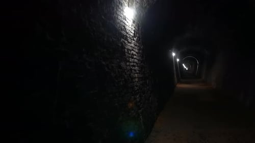 A dark tunnel with brick walls extends into the distance, illuminated by a series of lights mounted