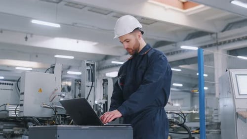 Using the laptop. Factory worker is indoors with hard hat.