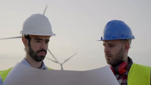 Close Up Two Engineers in Hard Hats Work Looking at Blueprint in Agricultural Field Wind Turbines