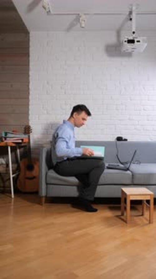 Vertical Video Young Man in a Blue Shirt and Dark Trousers Sits on a Gray Sofa in a Modern Living