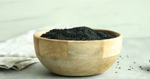 Pouring aromatic black caraway into wooden bowl at grey table, closeup