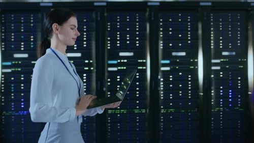 Beautiful Data Center Female IT Technician Walking Through Server Rack Corridor with a Laptop Compu