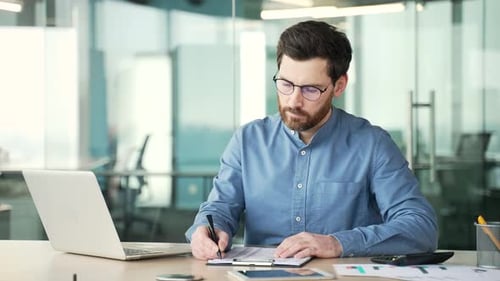 Busy entrepreneur in glasses is doing paperwork using a laptop computer while sitting at workplace.