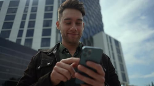 Lowangle View of Happy Young Man Smiling While Using Mobile Phone Standing on Background of Sleek