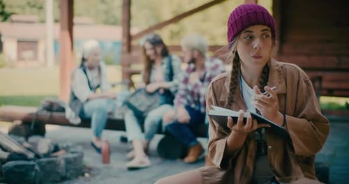 Young Woman Journaling Outdoors with Friends in Background