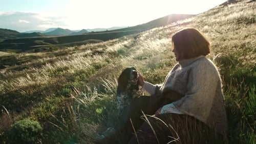 A Woman Caresses Her Dog Against the Backdrop of a Mountain Range in the Rays of the Sunset The