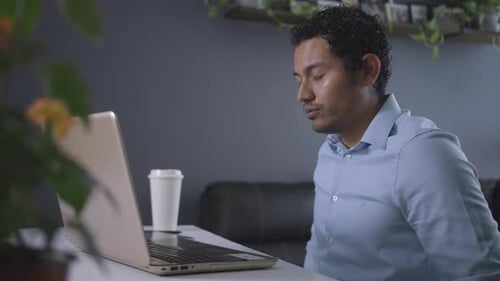 Hispanischer braunhäutiger Mann arbeitet am Laptop und trinkt Kaffee, während er müde ist
